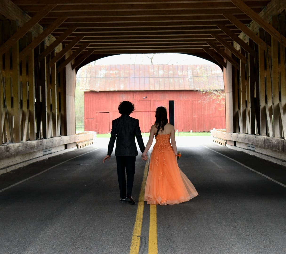 Couple walking hand in hand through a covered bridge toward a red barn