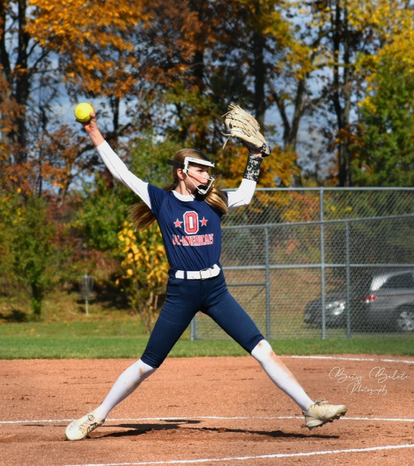 Softball pitcher mid windmill delivery on the mound with fall trees behind her