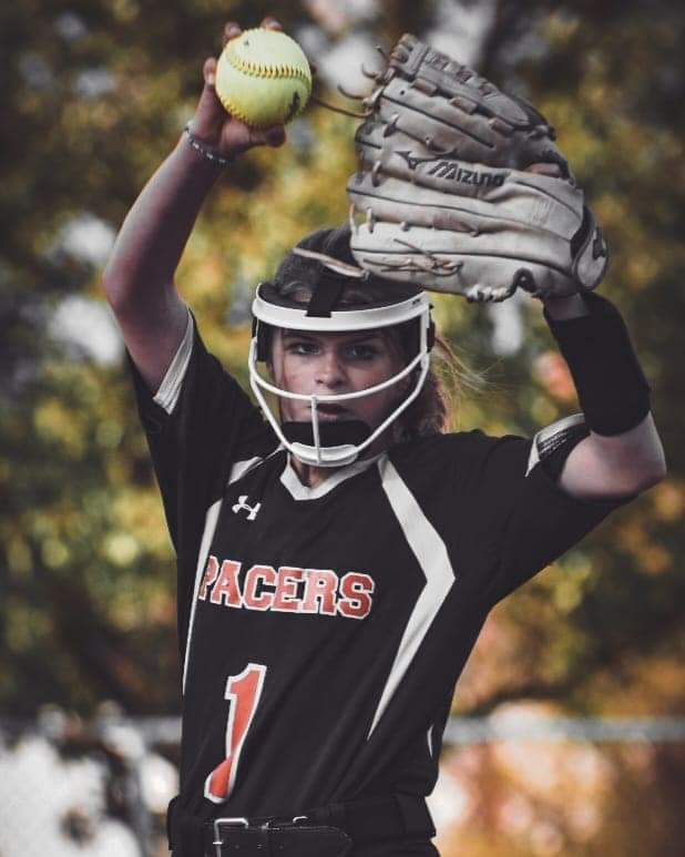 Fastpitch softball pitcher in black Pacers jersey at top of windmill motion with yellow softball raised high