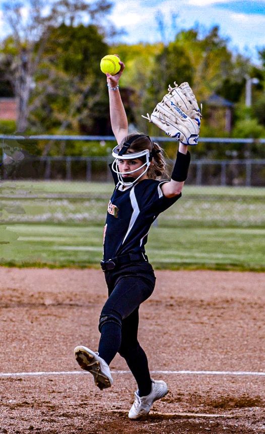 Fastpitch softball pitcher in navy uniform mid-windmill delivery with yellow softball raised high